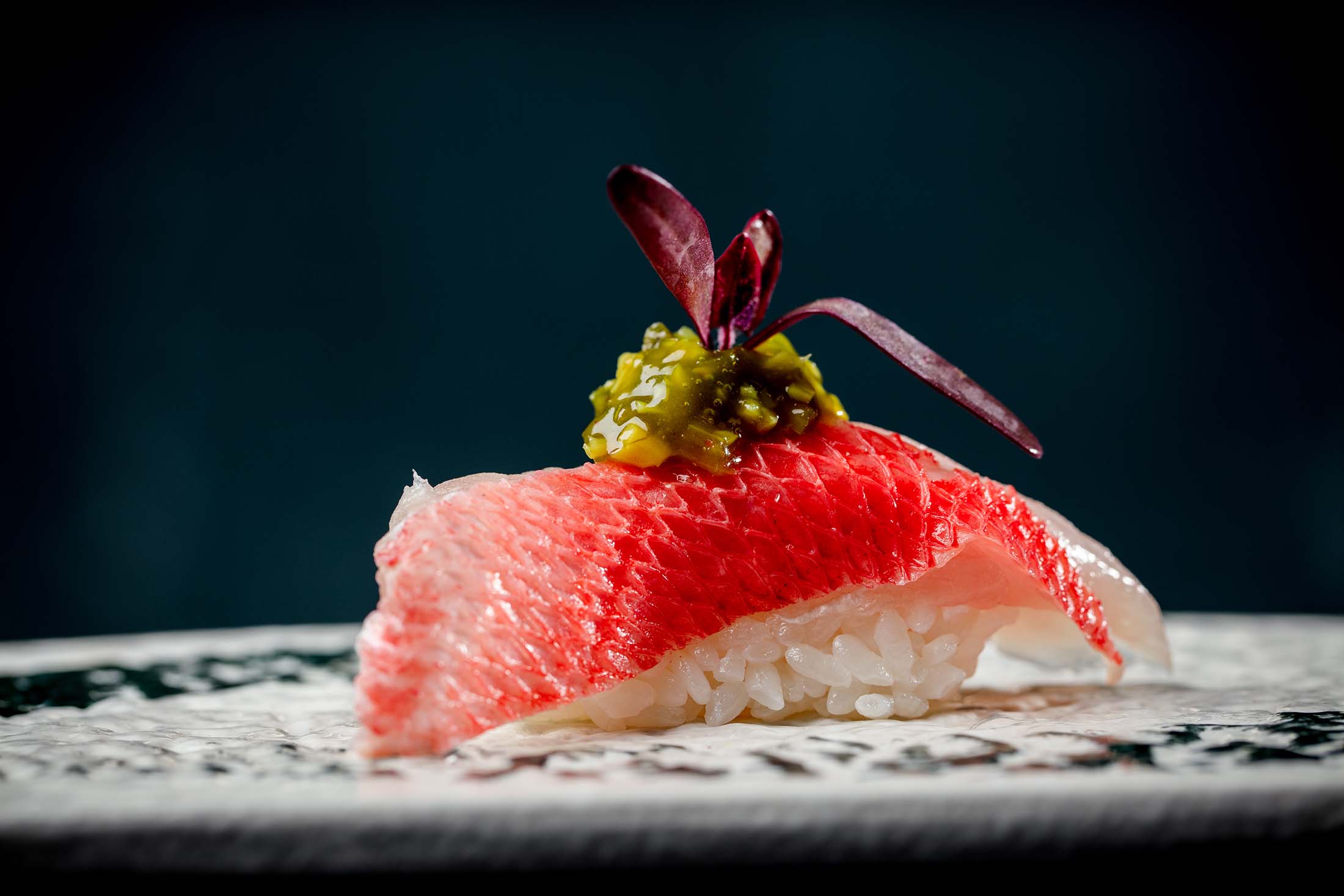 Nigiri sushi featuring a textured red fish slice topped with green relish and a microgreen