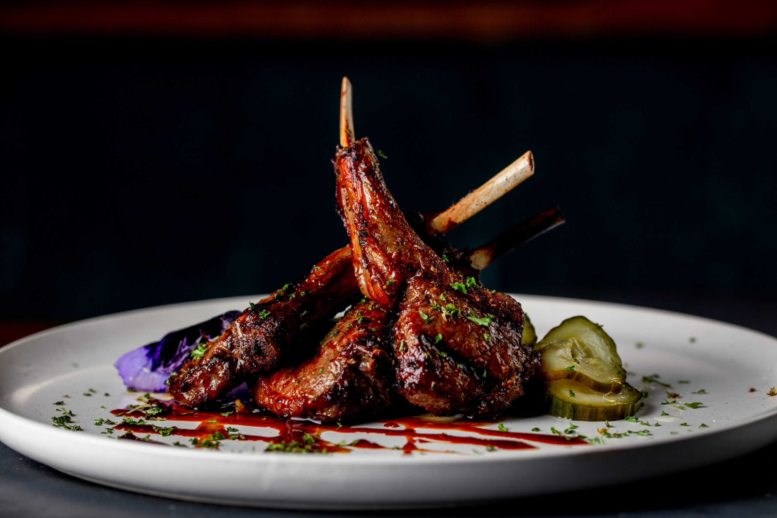 Three stacked grilled, glazed lamb chops on a white plate with sauce drizzle and seasoning