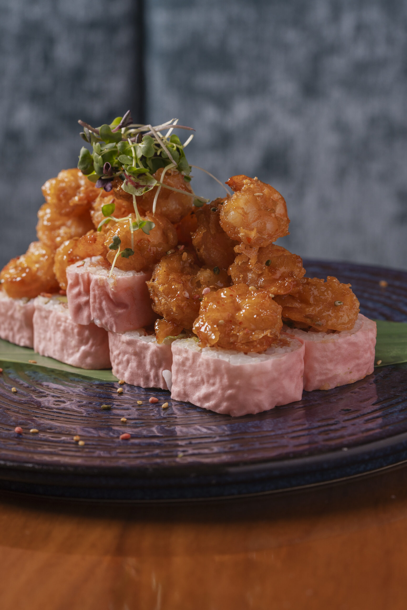Sushi roll with pink rice wrap topped with crispy glazed popcorn shrimp and garnished with microgreens