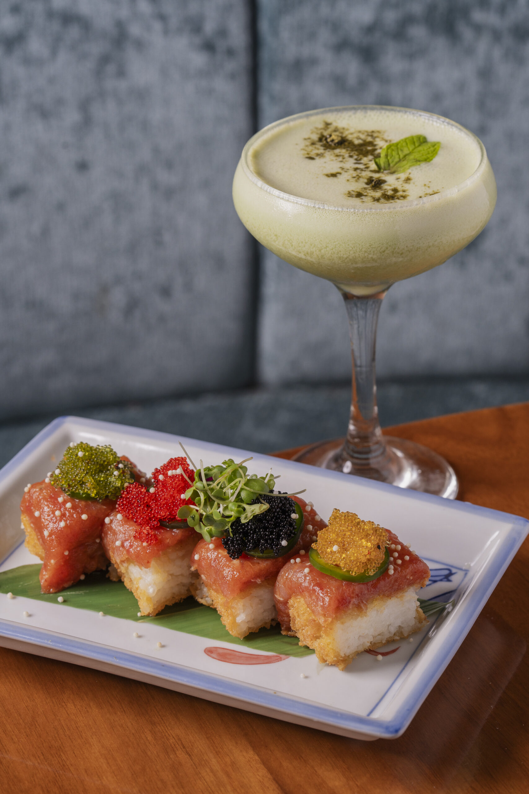 Four pieces of crispy rice with spicy tuna topped with assorted roe, microgreens, and jalapeño, served with a creamy green cocktail in a coupe glass.