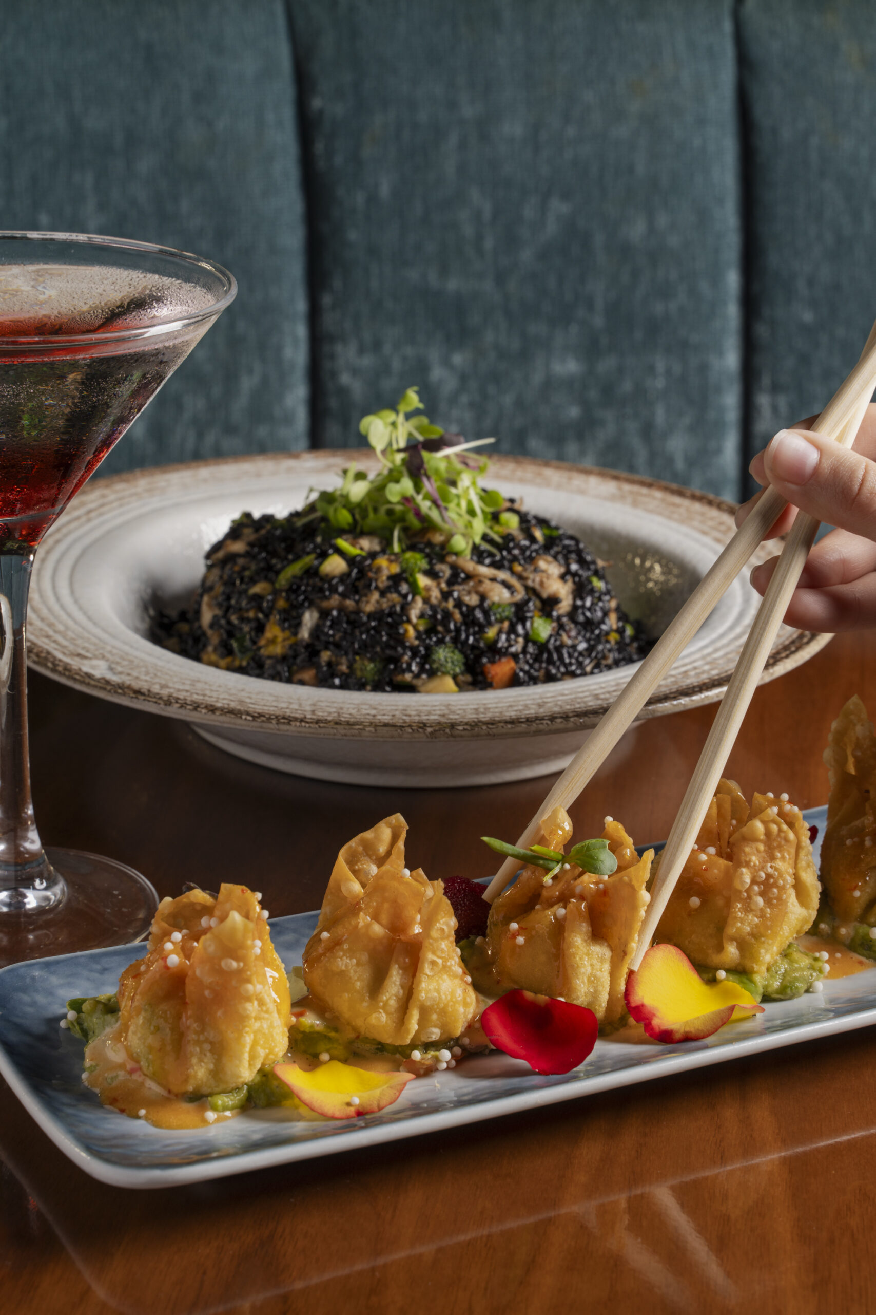 Golden-fried wontons garnished with edible flower petals on a plate, with a bowl of dark fried rice and a cocktail in the background